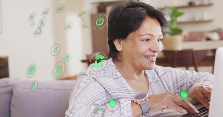 Typing senior Indian woman sitting on sofa in living room, with laptop, eco icons, copy space