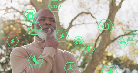 Standing man with gray beard holding chin in autumn park, with green ecology and energy icons