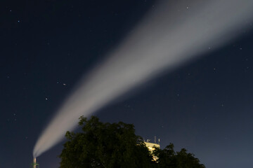 Smoke coming from factory chimney at night under starry sky with trees and building silhouette. Night industrial landscape photography. Air pollution and environment concept. Design for poster, banner