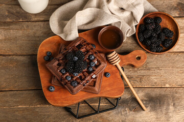 Board of sweet chocolate Belgian waffles with fresh blackberries, blueberries and bowl of honey on wooden background