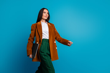Charming brunette businesswoman in retro-style outfit walking confidently with a laptop against a bright blue background