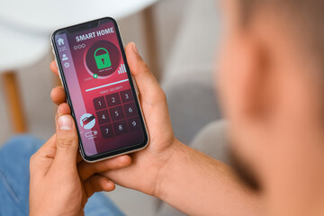 Young man using mobile phone with smart home security system application in room, closeup