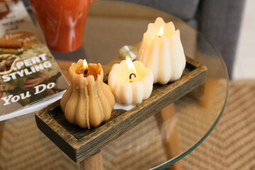 Wooden tray with burning candles in shape of pumpkins, magazine and tea cup on glass table at home, closeup