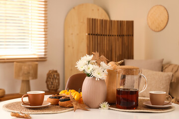 Table with hot tea, tasty poppy buns, flowers and autumn leaves on table in living room