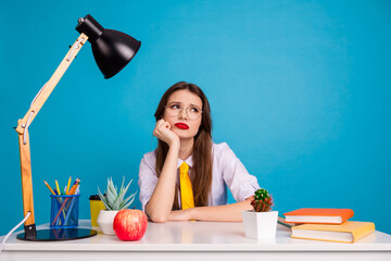Thoughtful young female student sitting at desk with book, lamp, and plants in front of a blue...