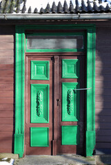 Decorative wooden door with green and brown carvings, set in a rustic wall with snow on the roof,...
