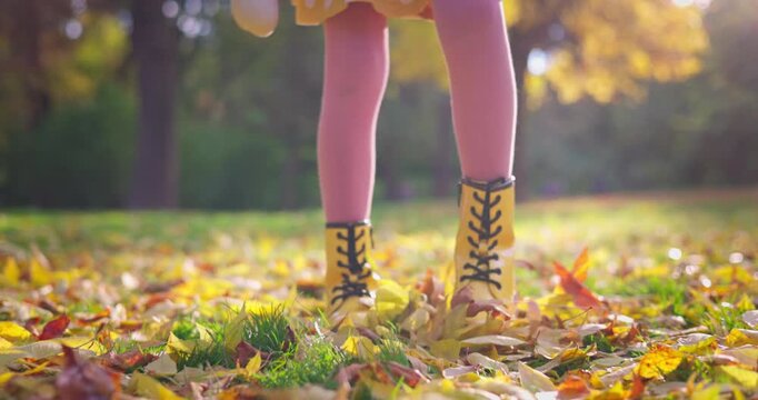 Little girl in yellow boots walking among falling autumn leaves in a sunlit park playing and enjoying fall season