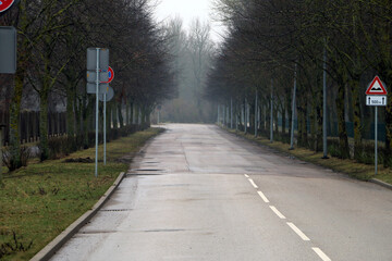 Empty wet road lined with bare trees on a misty day, featuring traffic signs and a warning sign for hazards ahead, evoking quiet, late autumn or winter atmosphere.