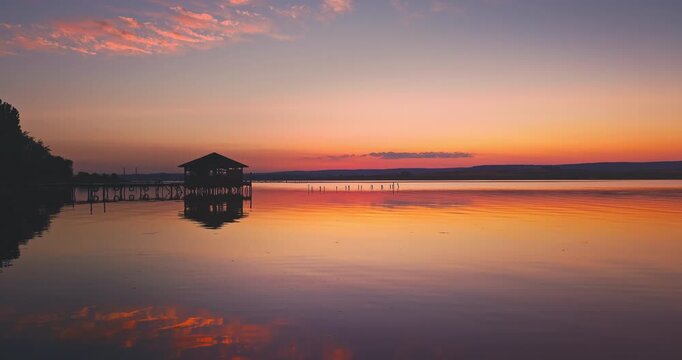 Scenic Sunset Over Wooden Fishing Jetty Pier With Gazebo Reflecting Golden Hour Colors and Calm Water Into Calm Lake At Dusk