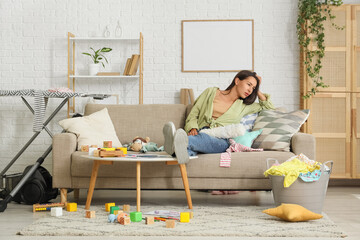 Tired mother with scattered toys and basket of laundry lying on sofa in messy living room