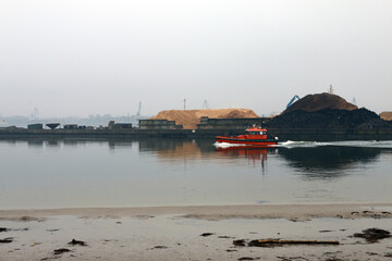 Industrial port scene with an orange boat on water, large piles of sand and coal on the dock, cranes in the foggy background, and a sandy shore with debris in the foreground.