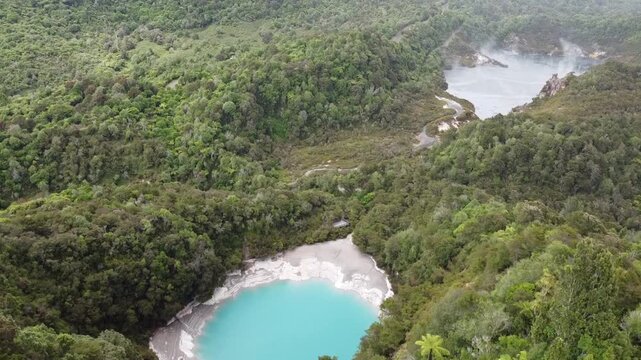 A drone captures a forward flight over Frying Pan Lake, a steaming geothermal hot spring in Waimangu Volcanic Valley, New Zealand. The shot shows geothermal steam, vibrant water, and lush green bush