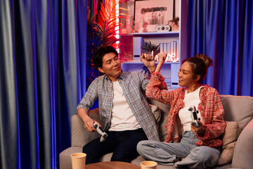 Couple gamer raising fist up holding joysticks be winner together complete level sitting sofa playing video game with front snack and drinks at neon light color living room at modern home. Infobahn.