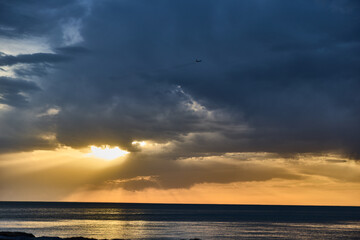 dramatic sunset ocean horizon with sun rays breaking through dark storm clouds over calm sea water and distant airplane silhouette