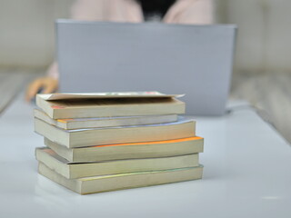 The image shows a stack of books on a table with a person and a laptop blurred in the background. This setting suggests a study or work environment.