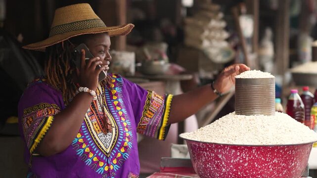 Small business owner, SME, Smiling African local traditional market woman in vibrant purple dashiki and straw hat talks on smartphone while selling grains at her outdoor market stall