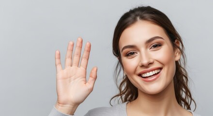 A young woman with brown hair smiles and waves hello with her right hand against a plain gray background.
