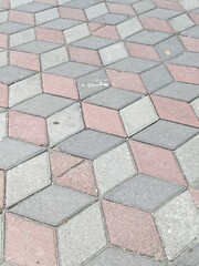 A fascinating outdoor pavement exhibits a striking three-dimensional optical illusion, formed by an intricate geometric pattern of grey and reddish-brown interlocking blocks.