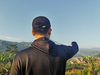 A person in a black hoodie and cap points towards a scenic mountain landscape under a clear blue sky.