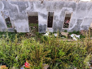 Dilapidated concrete wall with empty openings surrounded by dense, overgrown weeds and debris, suggesting an abandoned structure.