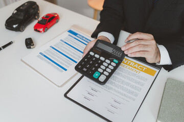 A businessman in a formal suit explains car insurance details, holding a small red car model and...