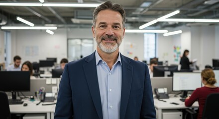 Obraz premium Smiling middle aged businessman in a dark suit jacket and light blue shirt stands confidently in a busy modern office environment
