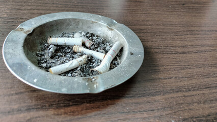 Closeup of an ashtray full of cigarette butts on a wooden textured table. Lots of dirty cigarette butts, butts, and filters in the ashtray. Copy Space for text. Selective focus.
