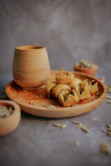 A small plate of handcrafted baklava sits next to a rustic wooden cup. The sweet pastries, filled with pistachios and nuts, are drizzled with a rich honey glaze. Scattered sunflower seeds add a touch 