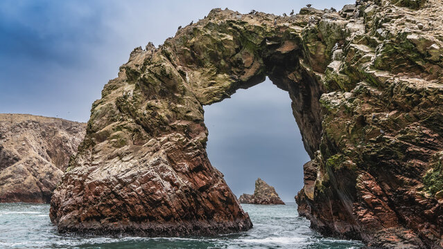 Picturesque natural stone arch over the ocean. Seabirds sit on the rocks. The islets are visible in the opening. Blue sky, clouds, turquoise sea. Paracas. Peru. Ballestas Islands