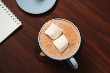 Hot chocolate in a blue cup with marshmallow and notepad on walnut table