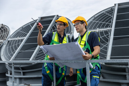 Two engineers in safety helmets and vests examine a construction blueprint on a rooftop HVAC site. Concept of industrial planning, project management, and mechanical system inspection. - Powered by Adobe