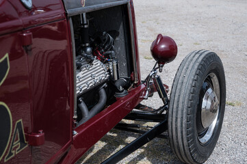 A close-up of the exposed V8 engine and front wheel of a classic American hot rod. The detailed shot highlights the mechanical parts and chrome suspension
