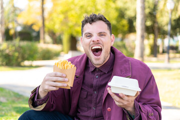 Young caucasian man at outdoors taking fried chips and takeaway box food with surprised expression