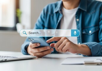 A person in a denim shirt is sitting at a desk with a laptop while holding and using a smartphone. A large, transparent search bar with a magnifying glass icon is floating in front of the phone, symbo