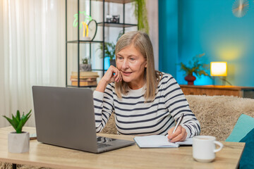 Fototapeta premium Mature woman participating in online course on laptop. Caucasian grandmother taking notes while sitting on sofa at home table, engaging in learning activities enhancing knowledge, education lessons