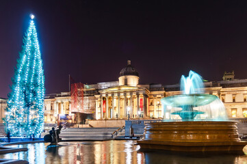 Long-exposure scene of London’s Trafalgar Square: the National Gallery’s classical facade glows behind a tall, blue-lit Christmas tree, while the foreground fountain creates soft water trails and refl