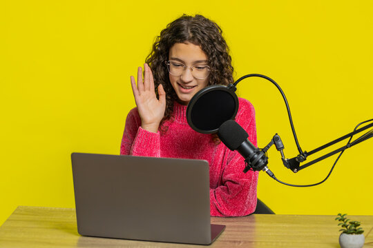 Happy excited blogger girl at table recording podcast giving workshop webinar. Young teen child influencer speaking into microphone and laptop webcam making video call discussing on yellow background - Powered by Adobe