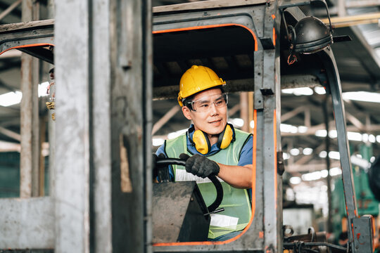 male worker asian is operating a forklift in a warehouse. He is wearing a blue shirt, a bright green safety vest, a yellow hard hat with ear protection around his neck, and clear safety glasses.