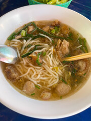 A bowl of noodle soup. The bowl contains white, thin rice noodles in a clear, brown broth. The noodles are topped with what appears to be sliced meat, meat balls, and chopped green onions.