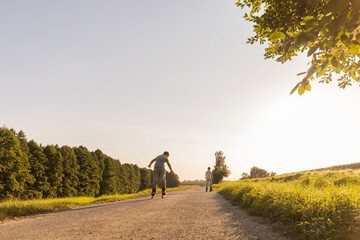 Obraz premium Two friends are inline skating along a path surrounded by greenery. The sun shines brightly, creating a joyful atmosphere for their outdoor activity