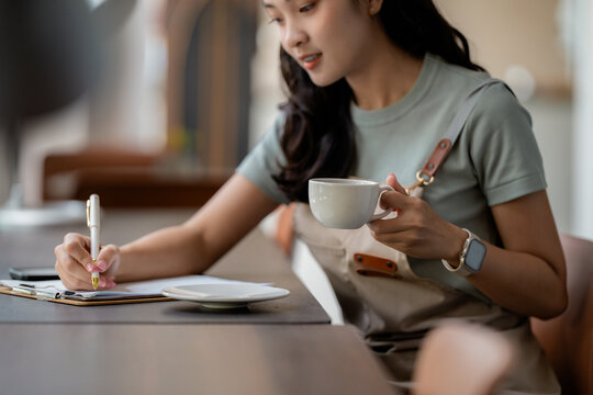 Woman barista entrepreneur planning her café: holding coffee cup, writing on clipboard with focused, professional expression