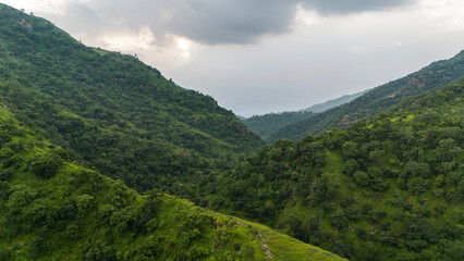Fototapeta premium Aerial photo of lush green Aravalli hills in Alsigarh, Udaipur, Rajasthan, during the monsoon season at evening, with dense trees, dramatic clouds in the sky, and the sun partially hidden behind them.