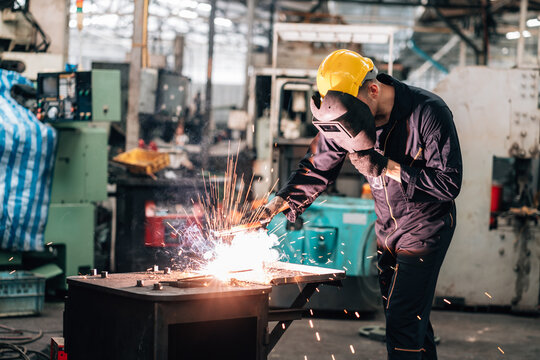worker is actively engaged in factory.wearing a dark blue jumpsuit and a yellow hard hat, with a welding helmet covering his face for protection. Sparks creating a bright flash of light with metal