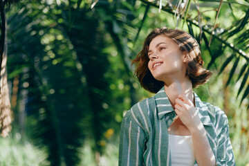 Fototapeta premium Portrait of a young woman outdoors, basking in sunlight amid lush greenery, wearing a striped green jacket and white top.