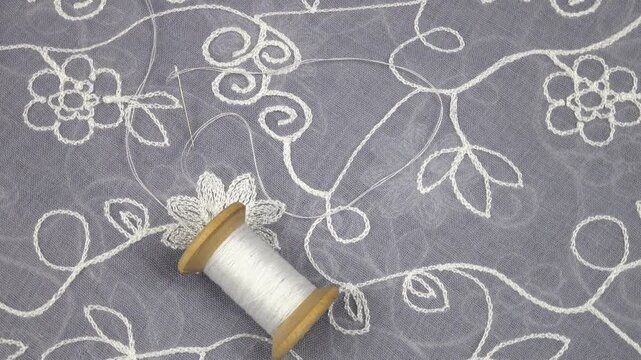 A spool of white thread and a needle and thread are on a gray- blue chiffon fabric with embroidered flowers, top view, rotation.  Fashionable clothing design elements, background