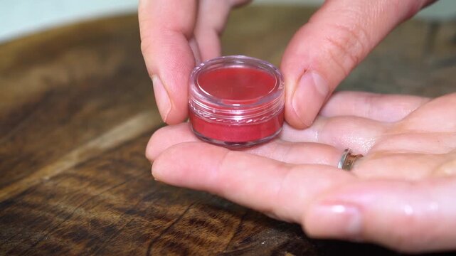 Making homemade natural lip balm with red pigment in transparent jar on dark wooden background. DIY beauty skincare concept with organic ingredients, eco-friendly cosmetics, and handcrafted style.