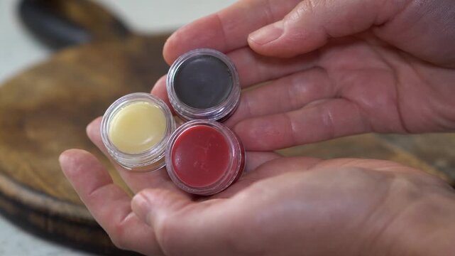 Woman&rsquo;s hands holding three natural homemade lip balms in clear containers. DIY cosmetics in red, brown, and transparent shades, organic skincare, eco-friendly beauty, and handmade products.