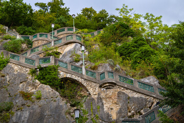 Naklejka premium Stone staircase with green railings winding up the rocky hillside surrounded by lush greenery. Picturesque architectural detail, perfect for travel, tourism, history, and exploration concepts.