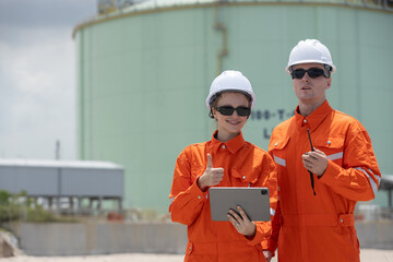 Two engineers in orange safety uniforms and helmets at an industrial gas plant. One holding a...