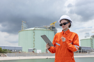 Female engineer in orange safety uniform and white helmet using a tablet while inspecting an industrial gas storage facility, representing technology, safety, and energy industry operations.